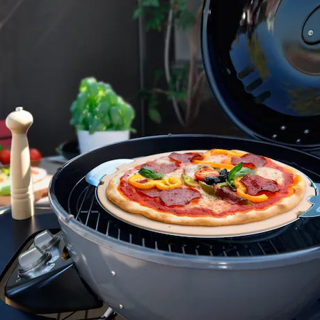 Eine Nahaufnahme einer Pizza, die auf einem Grill mit einem Pizzastein gebacken wird. Die Pizza ist mit Peperoni, Paprika und Basilikum belegt, und der Grill ist von Outdoor-Elementen wie einer Pfeffermühle und einer Basilikumpflanze umgeben.