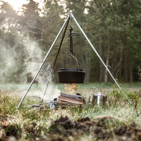 Ein Dutch Oven, der über einem offenen Feuer an einem Dreibein in einem Waldgebiet hängt.