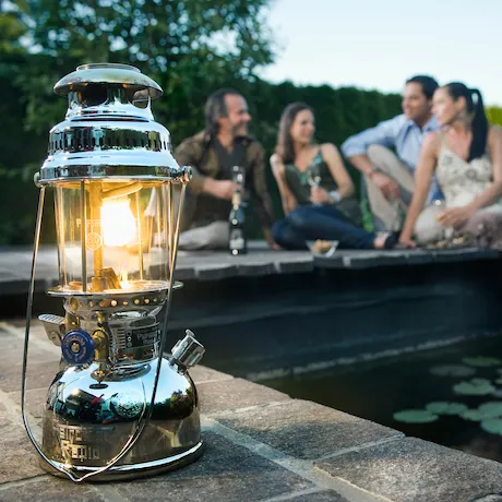 Eine glänzende Lampe steht im Freien auf einer Steinmauer, im Hintergrund sitzen Menschen und unterhalten sich