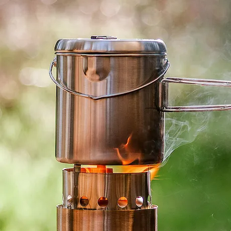 Ein Edelstahl-Kochgeschirr auf einem tragbaren Campingkocher, in dem ein Feuer brennt, mit einem verschwommenen grünen Hintergrund.