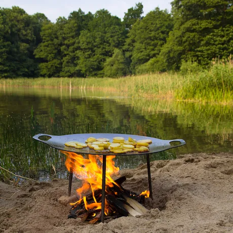 Ein Grill mit einem flachen, runden Kochfeld über offenem Feuer, aufgestellt am Ufer eines Sees mit üppiger Vegetation im Hintergrund.