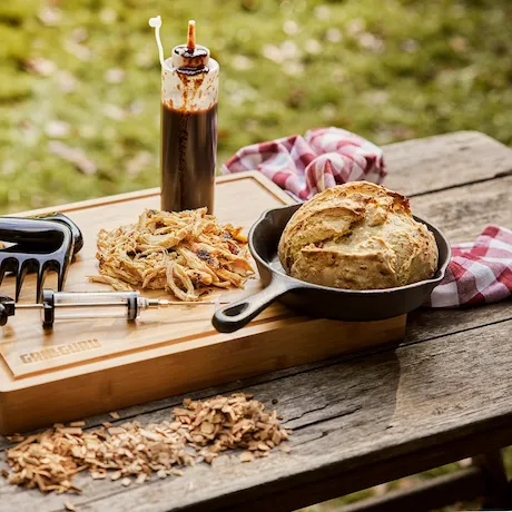 Schneidebrett mit gezupftem Fleisch, Soßenflasche, Brot und Grillwerkzeugen auf einem Holztisch