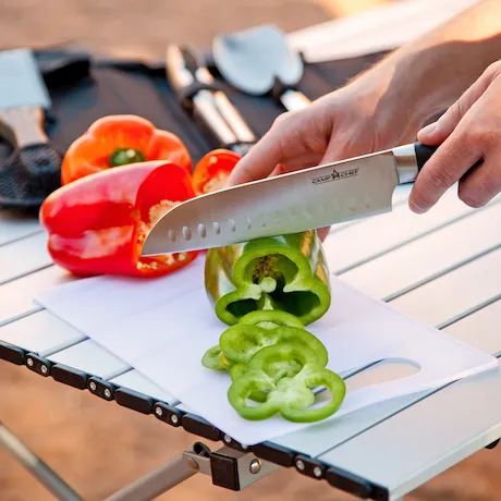 Hand, die grüne Paprika auf einem Schneidebrett mit einem Messer schneidet, im Hintergrund rote Paprika und Grillbesteck