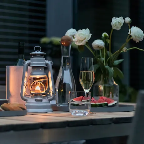 Ein gedeckter Tisch im Freien mit einer brennenden Laterne, einem Weinglas und Blumen im Hintergrund