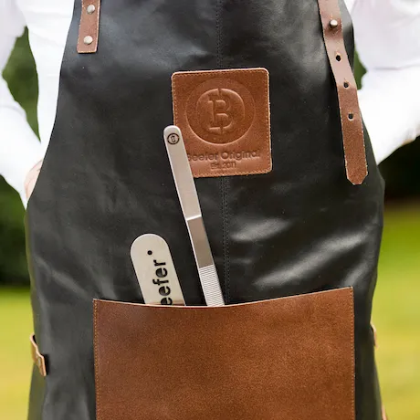 Eine schwarze Grillschürze aus Leder mit braunen Riemen und einer braunen Tasche. Auf der Schürze ist ein Lederpatch mit dem Logo und der Aufschrift 