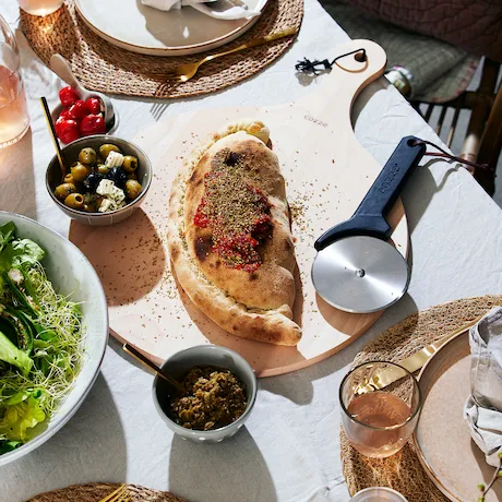 Gedeckter Tisch mit einem belegten Fladenbrot, Oliven, Salat und einem Pizzaschneider