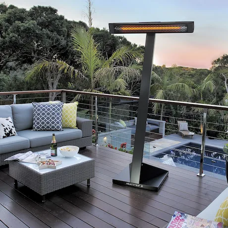 Ein gemütlicher Balkon mit Sitzbereich, Tisch und einem freistehenden Heizstrahler, im Hintergrund Palmen und ein Pool.