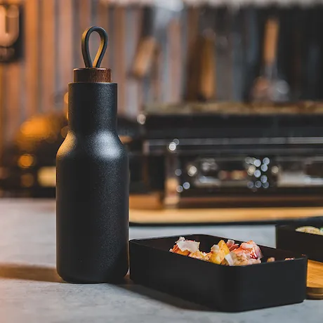 Eine schwarze Isolierflasche mit einer Holzabdeckung, die auf einem Tisch neben einer kleinen Box mit Gewürzen steht.