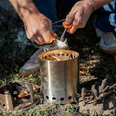 Hände, die einen Feuerstarter verwenden, um ein Feuer in einem tragbaren Edelstahlkocher im Freien zu entfachen.