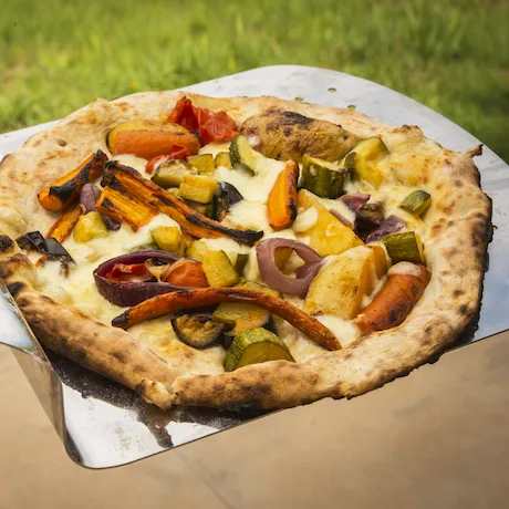 Eine frische Pizza mit gegrilltem Gemüse auf einem Pizzaschieber, mit einem Hintergrund aus Gras und einem Gehweg.