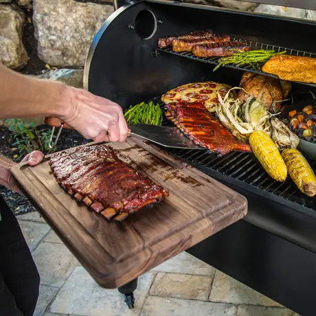 Eine Person, die Rippchen von einem Grillwagen auf ein Schneidebrett legt, mit verschiedenen gegrillten Lebensmitteln im Hintergrund