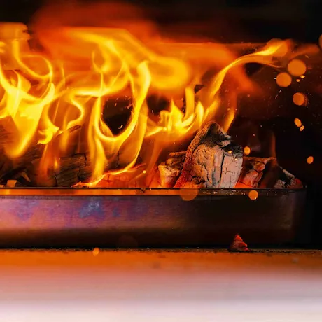 Nahaufnahme von brennenden Holzscheiten in einem Grill oder Ofen, mit intensiven Flammen und Funken.