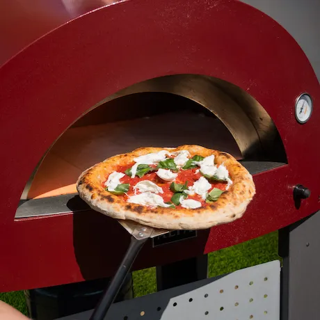  Eine Pizza, die mit einer Pizzaschaufel aus einem roten Outdoor-Pizzaofen herausgenommen wird. Die Pizza ist mit frischem Mozzarella, Tomatensoße und Basilikum belegt.