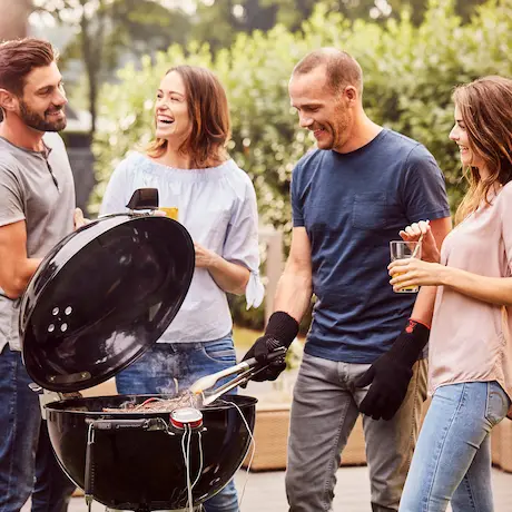 Eine Gruppe von vier Personen steht lachend um einen offenen Grill im Freien und bereitet gemeinsam Essen zu.