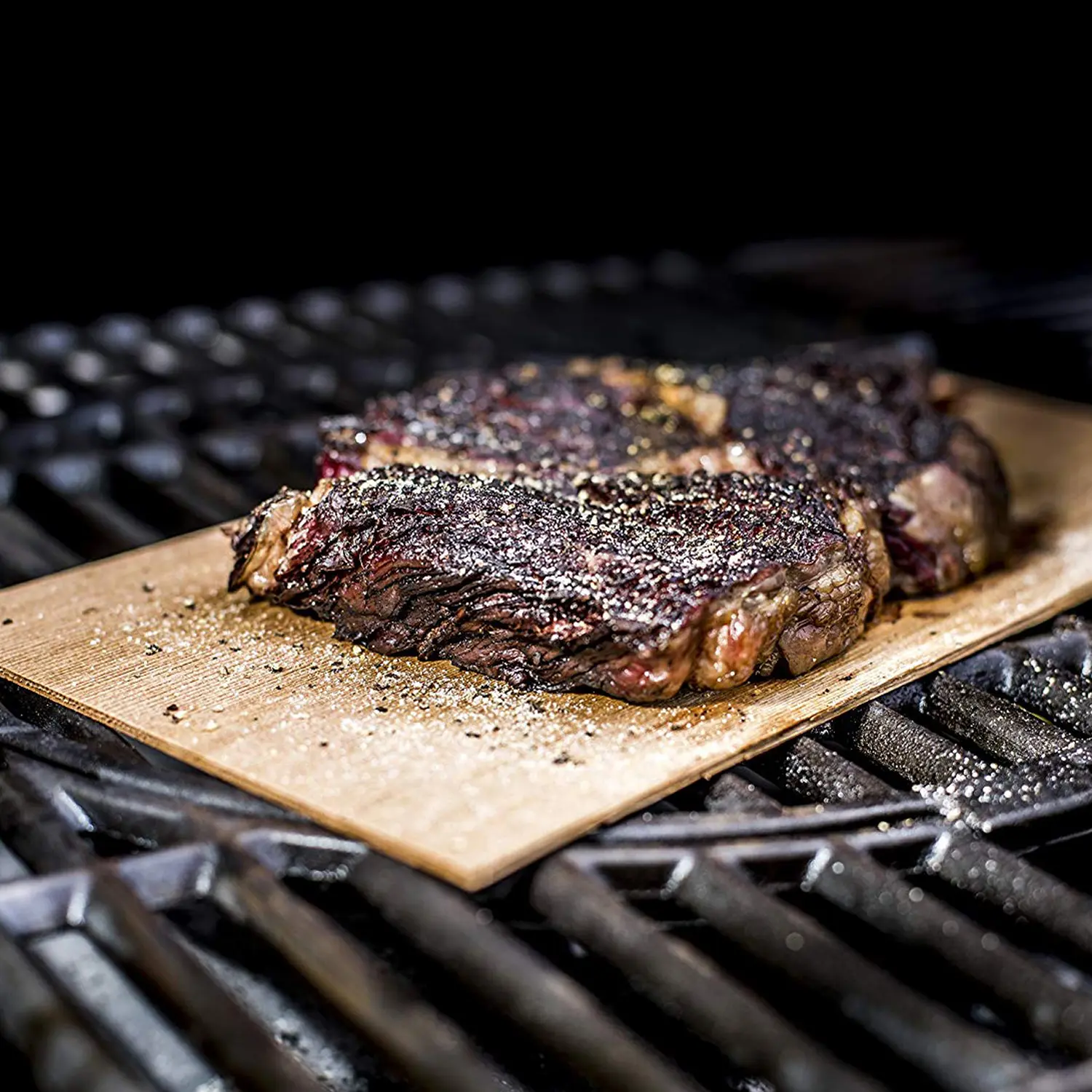 Zwei gebratene Steaks, gewürzt mit gemahlenem Pfeffer und Salz, werden auf einem Holzbrett über einem Metallgrill gegart.