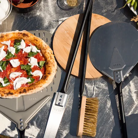  Eine Nahaufnahme einer Pizza auf einer Schaufel neben Pizzabackutensilien, darunter verschiedene Arten von Pizzaschaufeln und eine Reinigungsbürste. Die Pizza ist mit Mozzarella, Tomatensoße und frischem Basilikum belegt.