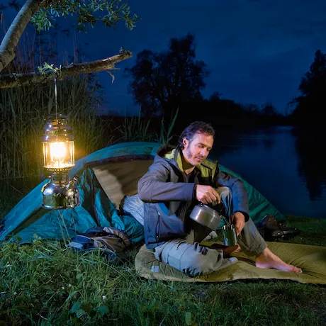 Mann beim Camping in der Nacht neben einem Zelt, hält eine Teekanne, mit einer hängenden Laterne im Hintergrund