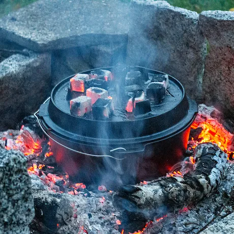 Dutch Oven mit glühenden Kohlen auf dem Deckel in einem Steinkreis mit brennenden Holzscheiten