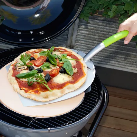 Eine Pizza wird mit einer Pizzaschaufel von einem Grill genommen. Die Pizza ist mit Tomatensoße, Oliven, Kirschtomaten und Rucola belegt.