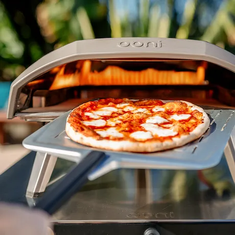 Eine frische Pizza wird mit einer Pizzaschaufel aus einem Ooni Pizzaofen geholt, der im Hintergrund brennt.